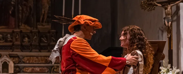 Due attori in scena davanti all'altare: lui, con il cappello arancione, le tiene le spalle e la guarda